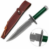 Rambo First Blood Standard Edition knife prop replica full hero view on mossy rock with brown leather RAMBO FIRST BLOOD embossed sheath snap button closure and leather tie lace showing grey silver satin display finish blade serrated saw-tooth spine green paracord-wrapped hollow handle and knurled silver pommel cap – movie display collectible