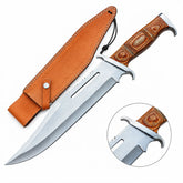 Rambo III Bowie knife prop replica held in gloved hand in sunlit green jungle forest showing silver satin display finish clip-point bowie blade with 2 elongated rectangular blade slots near ricasso and warm honey brown pakkawood handle with silver bolster panels – John Rambo 1988 movie display collectible