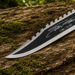 Rambo First Blood Part II Stallone Signature Edition knife prop replica dramatic elevated display on mossy log showing full black matte blade with serrated saw-tooth spine polished silver bevel black crossguard black diamond crosshatch knurled handle and knurled silver pommel cap in sunlit green jungle forest – Rambo fan gift wall display collectible