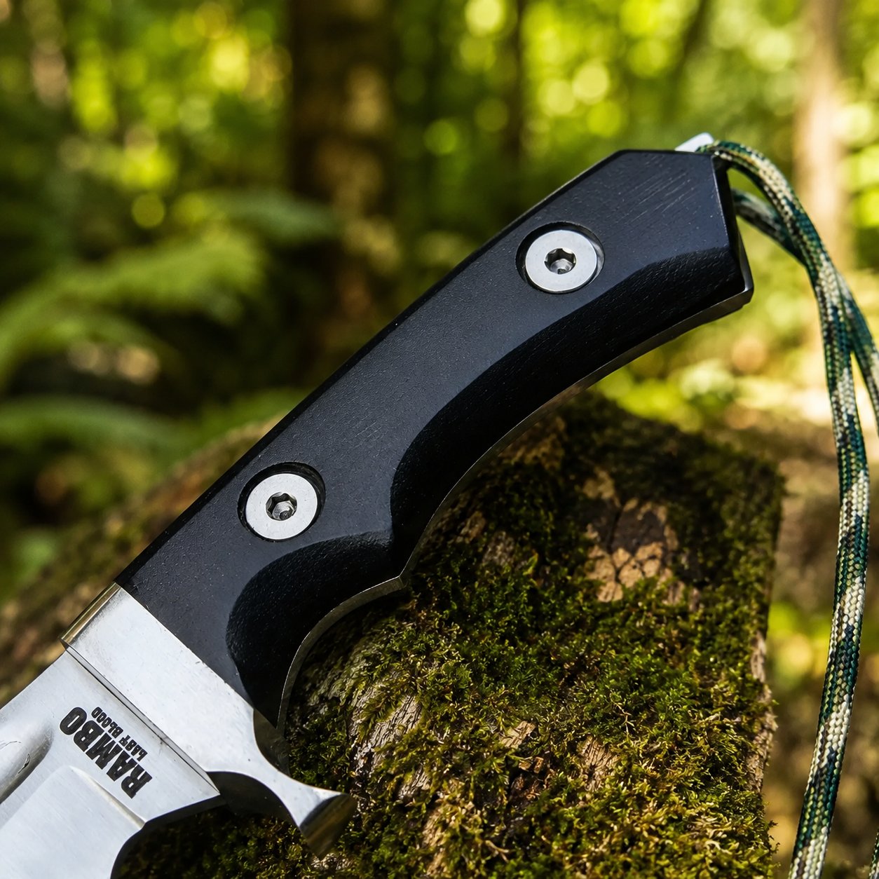 Rambo V Last Blood Bowie knife prop replica close-up macro of silver satin display finish clip-point bowie blade with single hollow grind RAMBO LAST BLOOD engraving small circular hole near ricasso and silver steel bolster guard with finger hook on mossy log in sunlit green forest – John Rambo Last Blood 2019 movie prop replica collectible