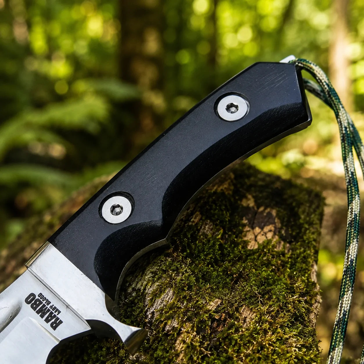 Rambo V Last Blood Bowie knife prop replica close-up macro of black G10 textured handle panels with smooth contoured grip 2 silver hex screw rivets and olive green camo paracord lanyard on mossy log in sunlit green forest bokeh – Rambo V Last Blood display collectible