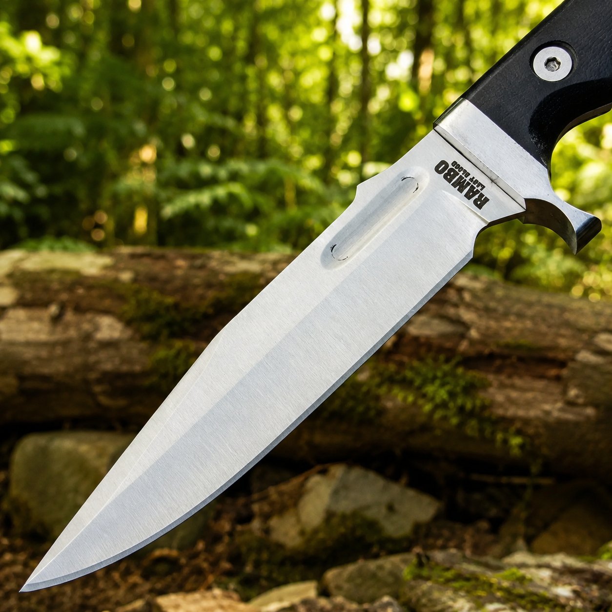 Rambo V Last Blood Bowie knife prop replica wide display view on mossy log showing full silver satin display finish clip-point bowie blade profile with RAMBO LAST BLOOD engraving black G10 textured handle silver steel bolster guard and olive green camo paracord lanyard in sunlit green forest – Rambo V Last Blood cosplay prop fan gift